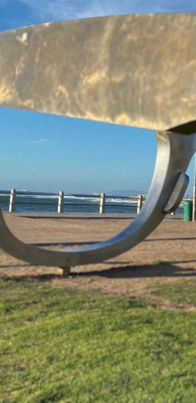 Brille Eine große, geschwungene Metallskulptur vor einem Strand mit Wellen im Hintergrund.