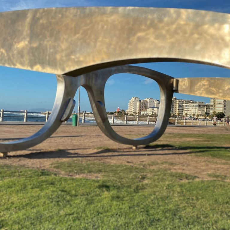 Große, stilisierte Brille aus Metall steht auf einer Wiese mit Stadtansicht im Hintergrund.